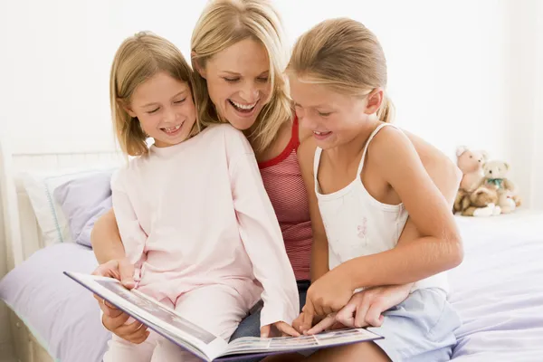 Woman and two young girls in bedroom reading book and smiling — Stock Photo, Image