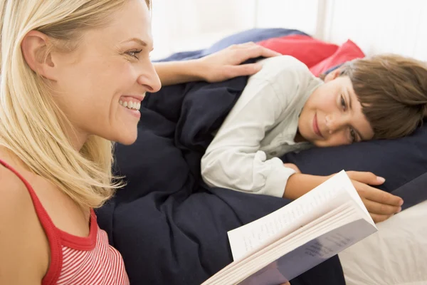 Donna che legge libro al giovane ragazzo a letto sorridente — Foto Stock