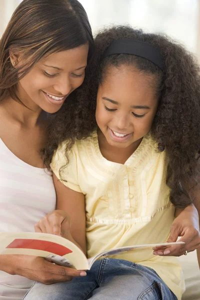Woman and young girl sitting in living room reading book and smi — Stock Photo, Image