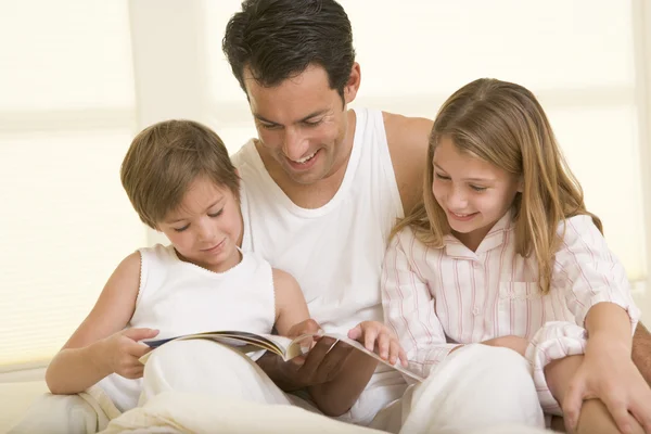 Мужчина с двумя маленькими детьми, сидя в кровати, читает книгу и смотрит телевизор — стоковое фото