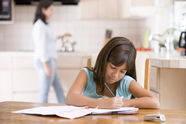 Молодая девушка на кухне делает домашнее задание с женщиной на заднем плане — стоковое фото