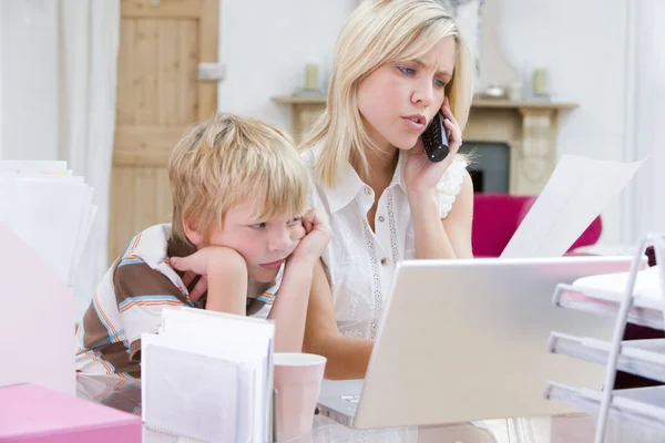 在家庭办公室用同时年轻男孩的笔记本电脑中使用电话的女人 — 图库照片