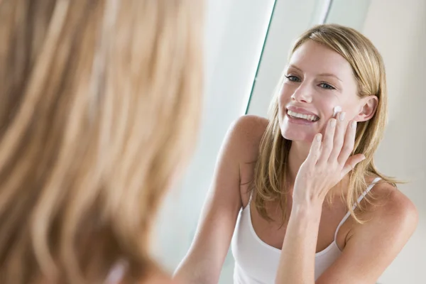 Женщина в ванной наносит крем для лица, улыбаясь — стоковое фото
