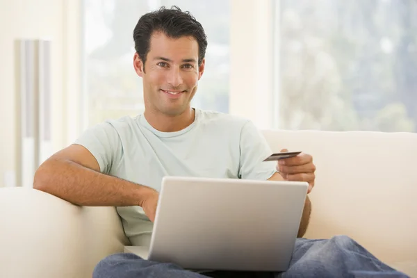 在客厅里使用的便携式计算机和持有信用卡微笑的男人 — 图库照片