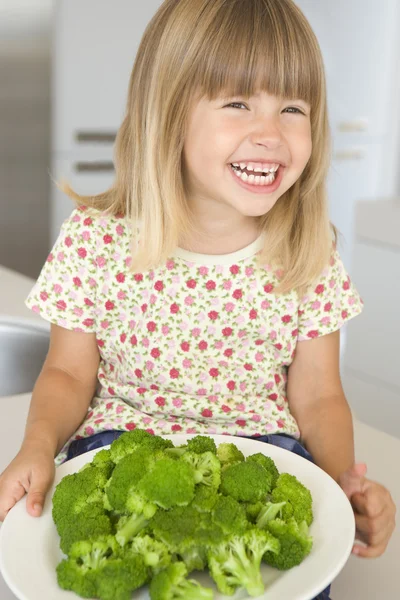 Νεαρή κοπέλα στην κουζίνα, τρώγοντας μπρόκολο χαμογελώντας — Φωτογραφία Αρχείου