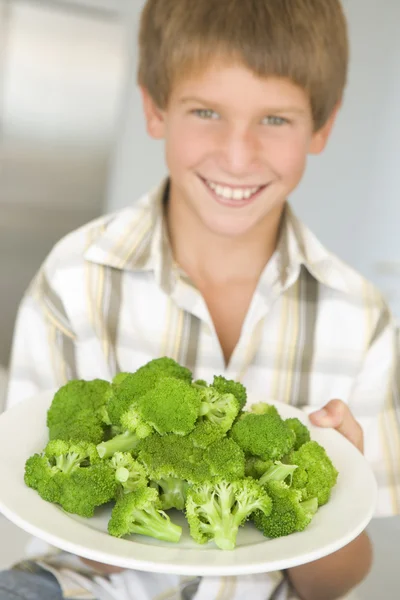 Young boy in kitchen eating broccoli smiling — Stock Photo, Image