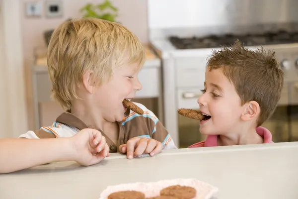Two young boys in kitchen eating cookies smiling — Stock Photo, Image