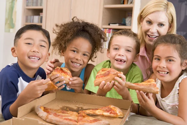Τέσσερα μικρά παιδιά σε εσωτερικούς χώρους με γυναίκα τρώει πίτσα χαμογελώντας — Φωτογραφία Αρχείου