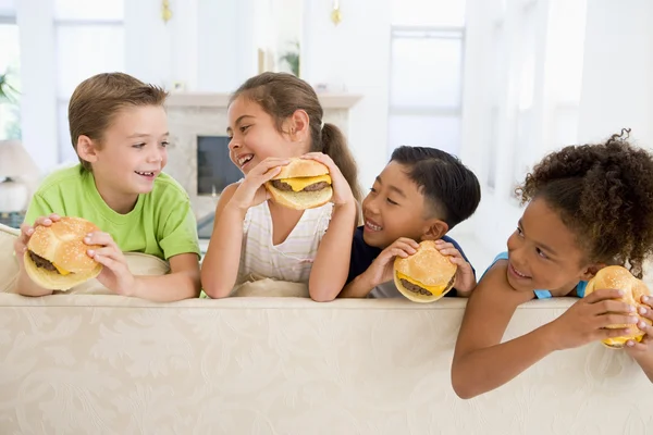 Quatro crianças pequenas comendo cheeseburgers na sala de estar sorrindo — Fotografia de Stock