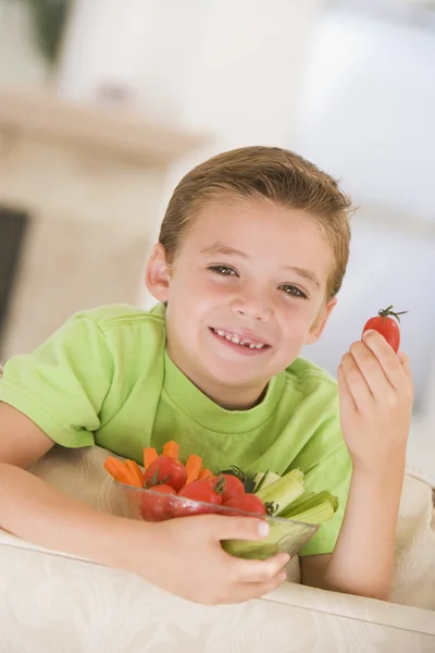 Ung pojke äta skål med grönsaker i vardagsrummet leende — Stockfoto
