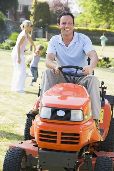 Man outdoors driving lawnmower smiling with family in background Stock Photo