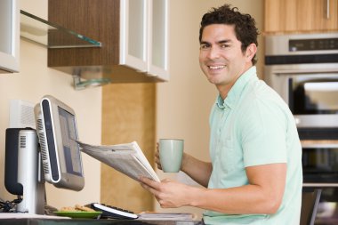 Man in kitchen with computer holding newspaper and coffee smilin clipart