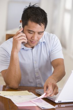 Man in dining room on cellular phone using laptop clipart