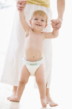 Mother helping baby walk indoors smiling clipart