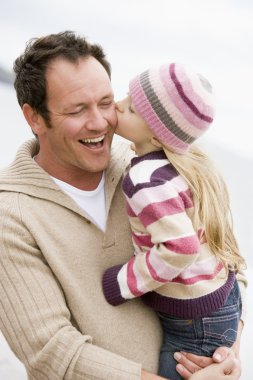 Father holding daughter kissing him at beach smiling clipart