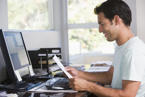 Homem em casa escritório usando computador segurando papelada e sorrindo — Fotografia de Stock