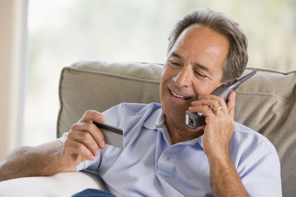Muž uvnitř pomocí telefonu a při pohledu na kreditní kartu s úsměvem — Stock fotografie