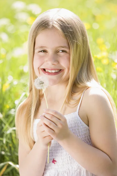 Chica joven sentada al aire libre sosteniendo la cabeza de diente de león sonriendo — Foto de Stock