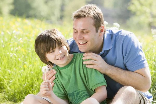 Far och son sitter utomhus med maskros huvud leende — Stockfoto