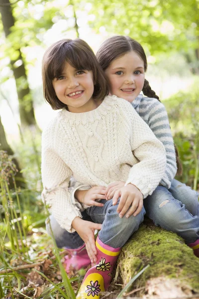 Two sisters outdoors in woods sitting on log smiling — Stock Photo, Image