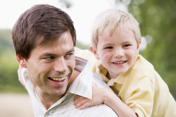 Pai dando filho piggyback passeio ao ar livre sorrindo — Fotografia de Stock
