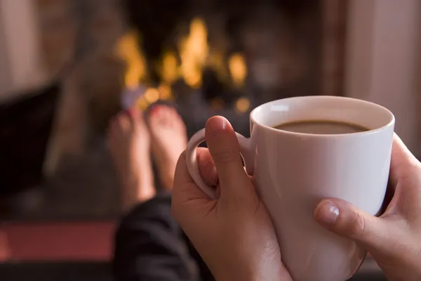 Feet warming at a fireplace with hands holding coffee — Stock Photo, Image