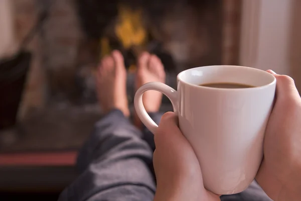 Pés de aquecimento na lareira com as mãos segurando café — Fotografia de Stock