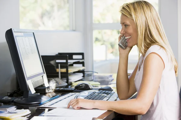 Жінка в домашньому офісі з комп'ютером за допомогою телефону посміхається — стокове фото