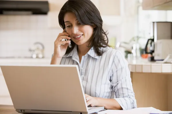 Kadın gülümseyerek cep telefonu kullanarak dizüstü bilgisayar ile mutfakta — Stok fotoğraf