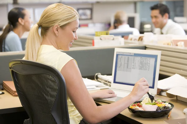Businesswoman in cubicle using laptop and eating salad — Stock Photo, Image
