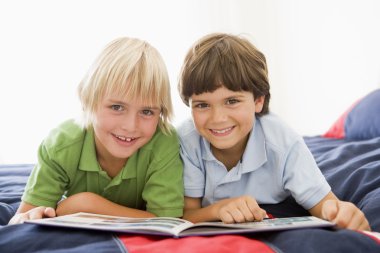 Two Young Boys Lying Down On A Bed Reading A Book clipart