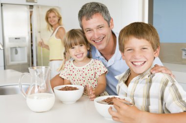 Father With Children As They Eat Breakfast And Mother In The Bac clipart