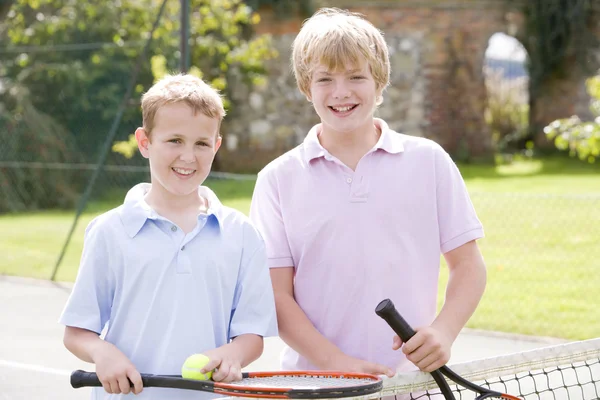 Two young male friends with rackets on tennis court smiling — Stock Photo, Image