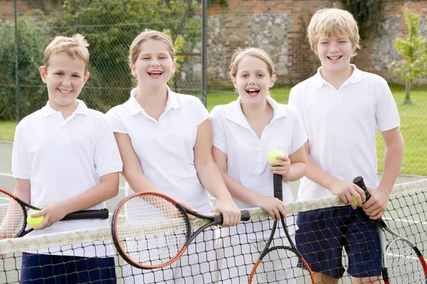 Fyra unga vänner med racketar på tennis court leende — Stockfoto
