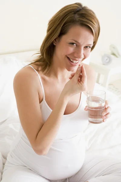 Беременная женщина в спальне принимает лекарства, держа воду улыбаясь — стоковое фото
