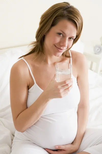 Hamile kadın gülümseyerek su cam ile yatak odası oturma — Stok fotoğraf