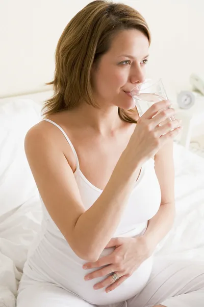 Mujer embarazada sentada en el dormitorio con un vaso de agua sonriendo —  Fotos de Stock
