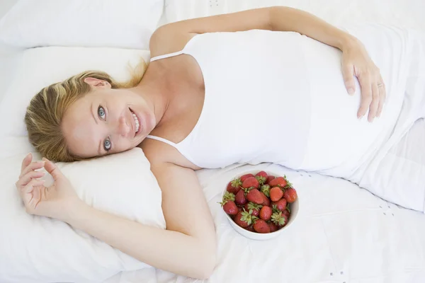 Pregnant woman lying in bed with bowl of strawberries smiling — Stock Photo, Image
