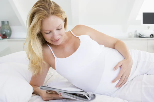 妊娠中の女性雑誌笑みを浮かべて読んでベッドで横になっています — ストック写真