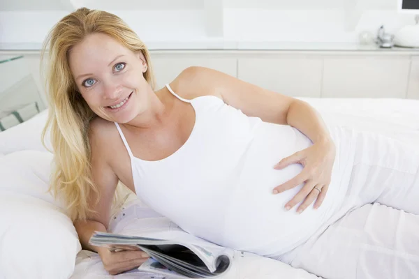 Беременная женщина лежит в постели читая журнал улыбаясь — стоковое фото