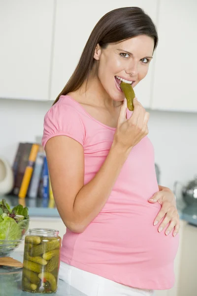 Zwangere vrouw in keuken eten augurken en glimlachen — Stockfoto