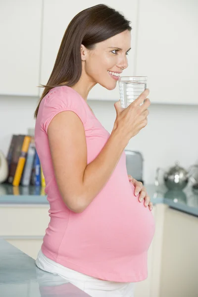 Mujer embarazada en cocina con vaso de agua sonriendo —  Fotos de Stock