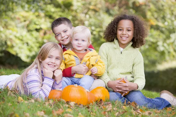 Three young friends with baby sitting on grass with pumpkins smi — Stock Photo, Image