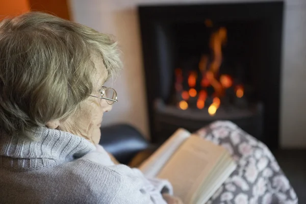 Starszy Kobieta Czytanie Książki Przez Ogień Domu — Zdjęcie stockowe