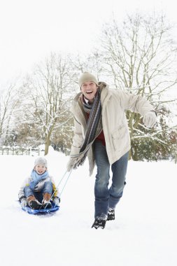 Father Pulling Son On Sledge Through Snowy Landscape clipart