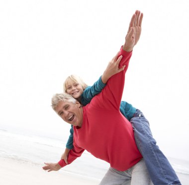 Grandfather Giving Grandson Piggy Back On Winter Beach clipart