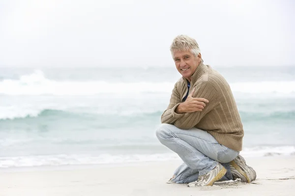 Hombre mayor en vacaciones arrodillándose en la playa de invierno — Foto de Stock