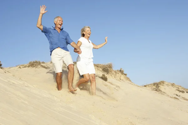 Casal sênior desfrutando de férias na praia correndo para baixo duna — Fotografia de Stock