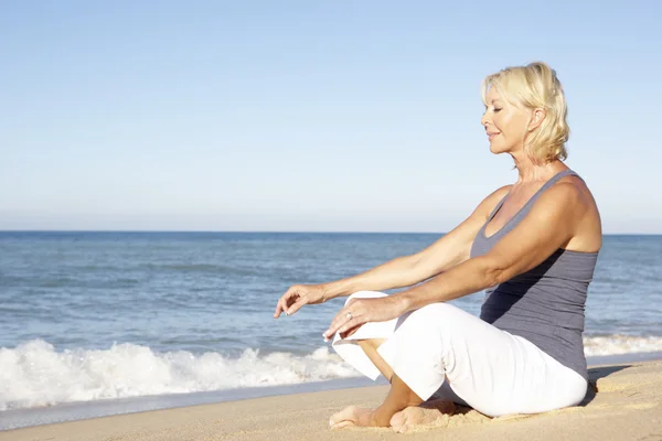 Senior vrouw in fitness kleding mediteren op strand — Stockfoto