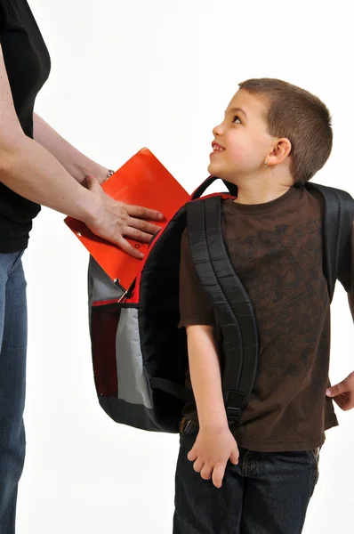 幸せな少年は 彼のバックパックにフォルダーを配置する Mom によって助けられています 彼は笑みを浮かべて お母さんを見て — ストック写真